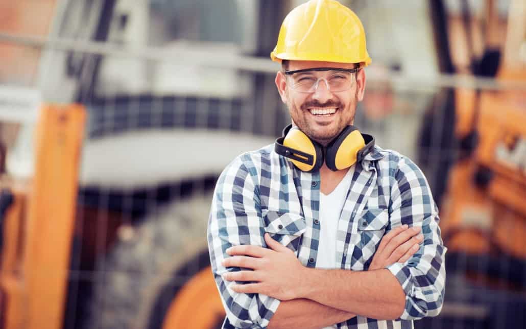 Smiling male construction worker wearing protective gear standing in front of industrial construction equipment
