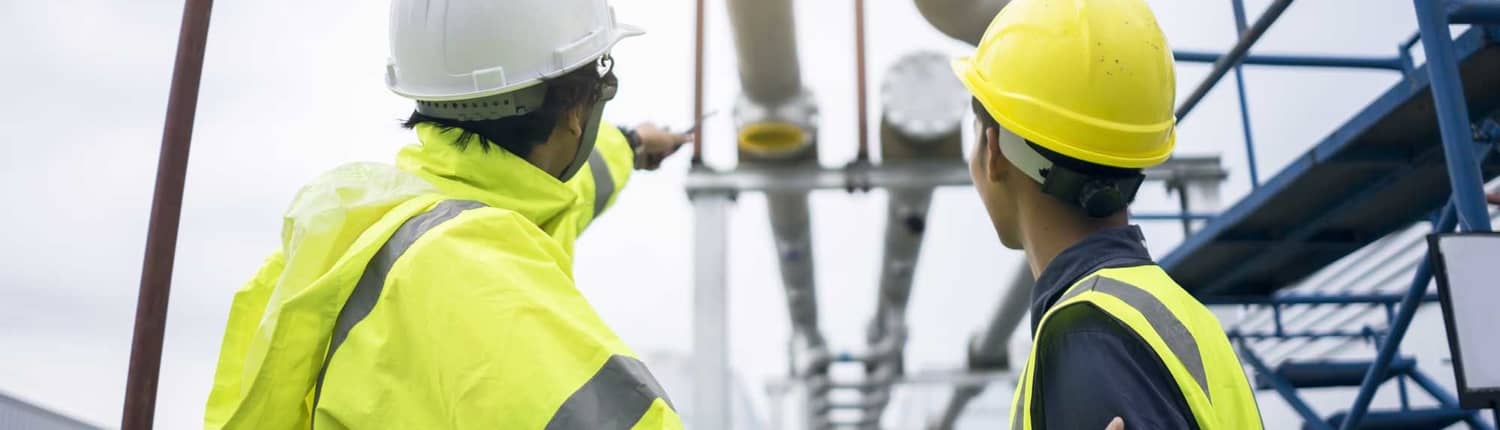 Two men in construction gear standing outside facing away from the camera looking up towards water piping system