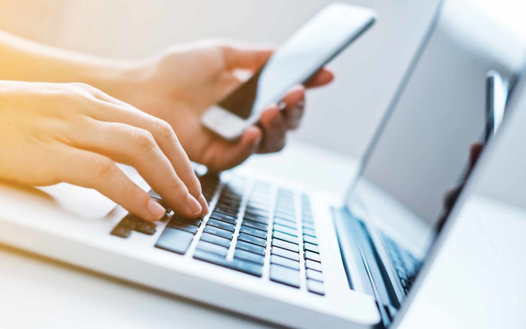 hands holding a smartphone and typing on a laptop